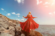 © svetograph - Red dress sea woman. A blonde with flowing hair in a long flowing red dress stands on a rock near the sea. Travel concept, photo session at sea