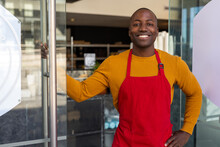 Portrait Of Happy African American Male Bakery Worker Wearing Red Apron At Open Door