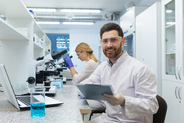 Portrait of a young male scientist looking into the camera with a smile. He is sitting in the laboratory in front of a microscope with a documents, a female colleague in the background.