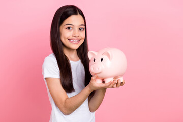 Wall Mural - Photo portrait of pretty young child girl hold piggy bank collect education dressed stylish white clothes isolated on pink color background