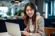 © Surachetsh - Asian office working girl with a radiant smile sits in front of her laptop computer, immersed in work, against a soothing blue turquoise background. generative AI.