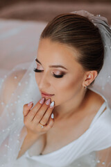 Wall Mural - A gorgeous bride with a classic hairstyle, wearing a long robe and a long veil, is sitting and posing on a white sofa in her room. The bride on a white background