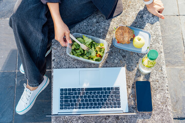 Wall Mural - Top view business woman eating salad from lunch box sitting on the bench at office park. Balanced diet lunchbox with sandwich, apple and water. Healthy eating habits and well-being. Selective focus