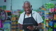 © Marco - Happy Portrait of Brazilian Senior Supermarket Staff Holding Tablet, Joyful Older Male Employee in Apron
