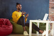 © master1305 - Young african man sitting on floor pillow, talking on mobile phone with friends and colleagues. Lively discussion. Concept of business and education, freelance job, modern lifestyle