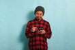 Happy young Asian man wearing eyeglasses, a beanie hat, and a red plaid flannel shirt is reading chat or news on his smartphone, isolated on a blue background