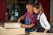 © EKKAPON - Senior male carpenter instructing young woman to use equipment while working at carpentry workshop.