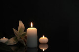White lily and burning candles on black mirror surface in darkness, space for text. Funeral symbols