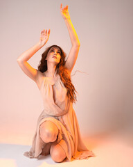 Wall Mural - Full length portrait of beautiful brunette  female model wearing dress with golden back light lighting. Kneeling pose with gestural arm pose.  isolated on studio background.