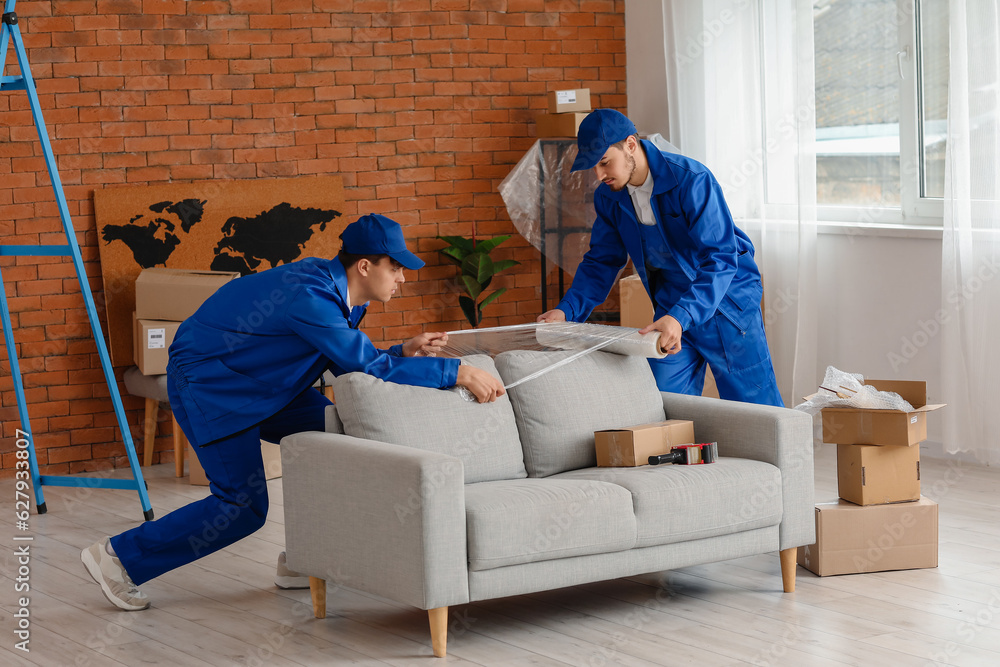 Male movers wrapping sofa with stretch film in room