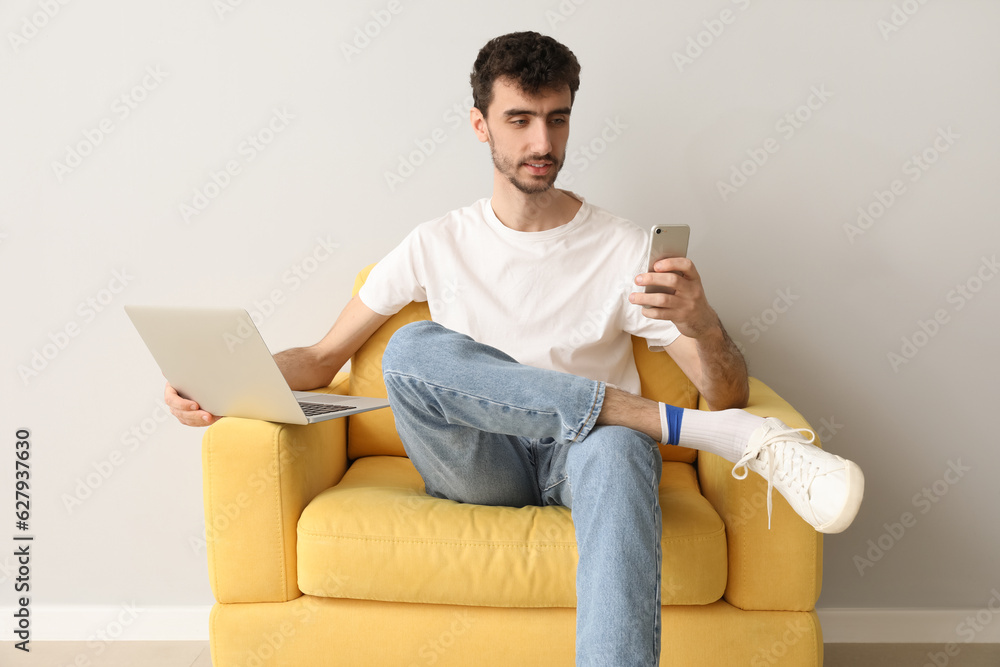 Young man with laptop using mobile phone in armchair near light wall