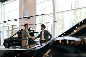 Wall Mural - Remote view of young customer male thanking stylish dealer male wearing suit and eyeglasses for helping to choose car to buy in dealership center. Concept of choosing and buying new car at showroom