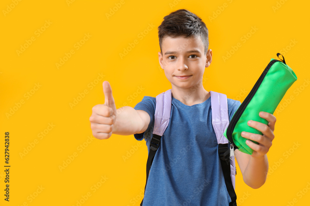 Little schoolboy with pencil case showing thumb-up on orange background