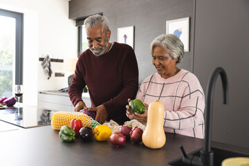 Happy senior biracial couple unpacking bags of vegetables in kitchen at home