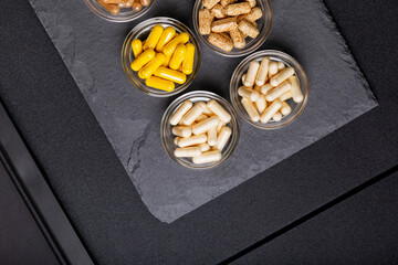 Wall Mural - Various vitamins and minerals, food supplements in forms of capsules in small glass bowls from above on blackstone desk on black background. Nutritional supplements to improve health. Healthy life.