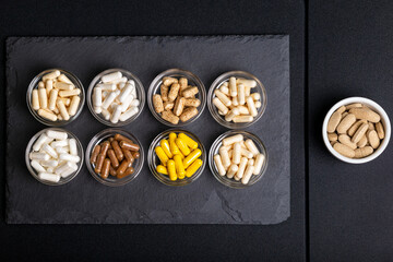 Wall Mural - Various vitamins and minerals, food supplements in forms of capsules in small glass bowls from above on blackstone desk on black background. Nutritional supplements to improve health. Healthy life.