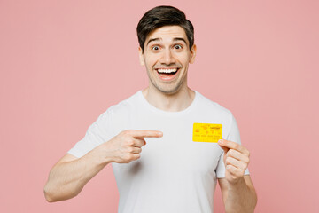 Poster - Young smiling happy man wearing white t-shirt casual clothes hold in hand point finger on mock up of credit bank card isolated on plain pastel light pink background studio portrait. Lifestyle concept.