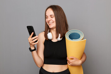 Smiling happy woman holding rolled fitness mat dressed in activewear isolated over gray background using modern mobile phone downloads application for sport training.