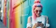 © Zaleman - A hipster teenage girl with pink pigtails uses a smartphone on the background of a multi-colored street wall.