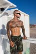 Chico joven musculoso y tatuado posando sin camiseta y en bañador frente a chiringuito de playa