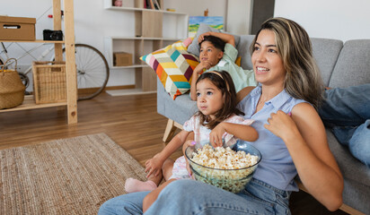 Wall Mural - Beautiful young Hispanic mother and her children are watching TV, eating popcorn and smiling while sitting on the couch at home