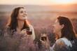 Two happy women drinking healthy herbal tea, sitting in a beautiful lavender field at sunset.