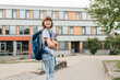 © Elena Medoks - Portrait of a happy teenage girl standing with a backpack on her back in front of the school with textbooks in her hands. Back to school, start of the school year.