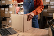 Worker man In Retail Warehouse working packing parcel box with tape dispenser for shipment. Delivery and distribution center full of shelves with goods. Close-up