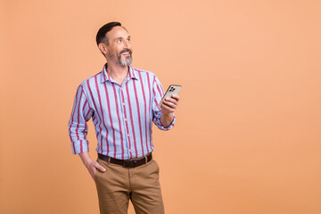 Poster - Photo of pleasant friendly man wear striped shirt brown trousers look at empty space hold smartphone isolated on beige color background