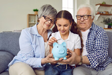 Loving Grandparents Help Granddaughter Save Up Money In Piggy Bank. Happy, Smiling Child Girl Together With Grandma And Grandpa Sitting On Sofa At Home And Putting Coins In Family Piggybank Moneybox