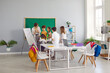 © Studio Romantic - Happy children at private school take turns answering tasks of young teacher near the blackboard. Cool school room in modern style with large communal table and chairs with hanging bright backpacks.