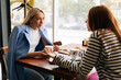 © dikushin - Side view of two pretty young women friends sitting at coffee shop having coffee and chatting sitting at table by window. Happy redhead and blonde female friends meeting at cafe on weekend.