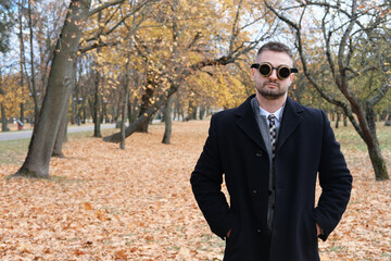 A man in a business suit, coat and steampunk goggles poses in an autumn park