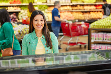 Female customer smiling looking into display case of bakery counter in grocery store, customers in produce section in background 