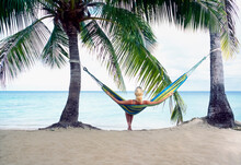  Woman Sitting In Hammock Tied Between Two Palm Trees In Tropical Paradise