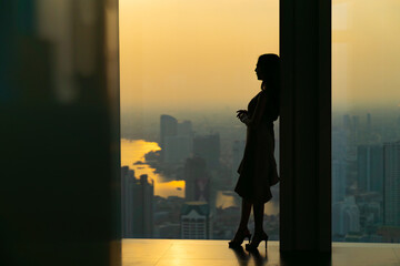 Silhouette Asian woman standing by the window at skyscraper building in metropolis and looking cityscape at summer sunset. Beautiful girl enjoy urban lifestyle activity in the city on holiday vacation