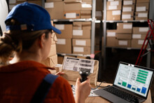 Woman Worker Working With Warehouse Management System With Barcode Reader And Laptop Computer. Girl Working In Factory Warehouse Scanning Labels On The Boxes With Barcode Scanner.