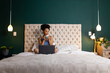 © Wavebreak Media - Biracial woman sitting on bed, drinking coffee and using laptop in bedroom, copy space