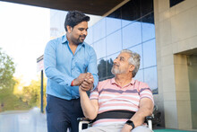 Young Man Giving Support To Sick Old Man In A Wheel Chair.