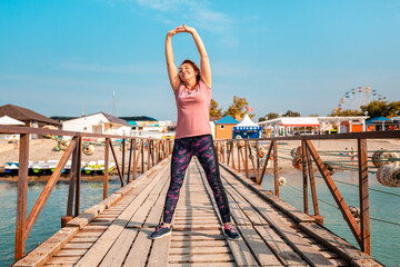 Summer activities. An adult happy caucasian woman in sports clothes is doing sports on the pier by the sea. Sports and healthy lifestyle