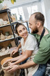 © LIGHTFIELD STUDIOS - Joyful craftswoman in apron looking at boyfriend and molding clay on pottery wheel in workshop