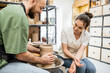 © LIGHTFIELD STUDIOS - Smiling craftswoman in apron sitting near boyfriend making clay vase on pottery wheel in workshop