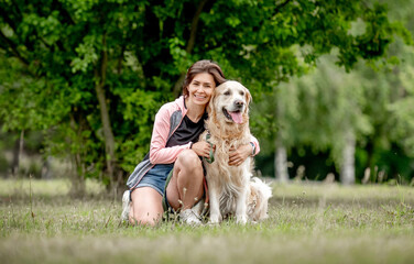 Wall Mural - Pretty girl with golden retriever dog sitting at nature. Beautiful young woman hugging purebred pet doggy labrador in park at summer