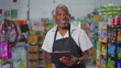 © Marco - Joyful Happy Brazilian senior employee of supermarkert wearing apron and holding tablet device, portrait of a black older male staff depicting job occupation at retail store