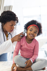 african american female doctor examining girl patient using otoscopy at hospital