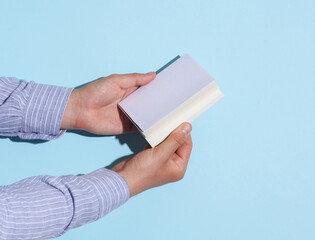 Man's hands in shirt holding book with white blank cover on blue background with shadow