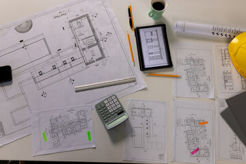Overhead view of desk with architectural blueprints, tablet, coffee and hardhat