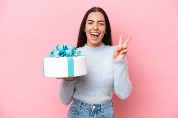 Poster - Young caucasian woman holding birthday cake isolated on pink background smiling and showing victory sign