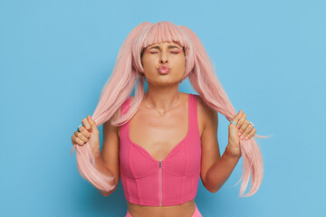Wall Mural - Doll style girl in pink top posing on blue background, holding her long pink ponytails in hands and sends airkiss, barby trend concept, copy space