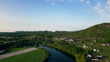 Canvas Print - Drone footage over rural countryside houses on lawn by the river with green hills in Norway